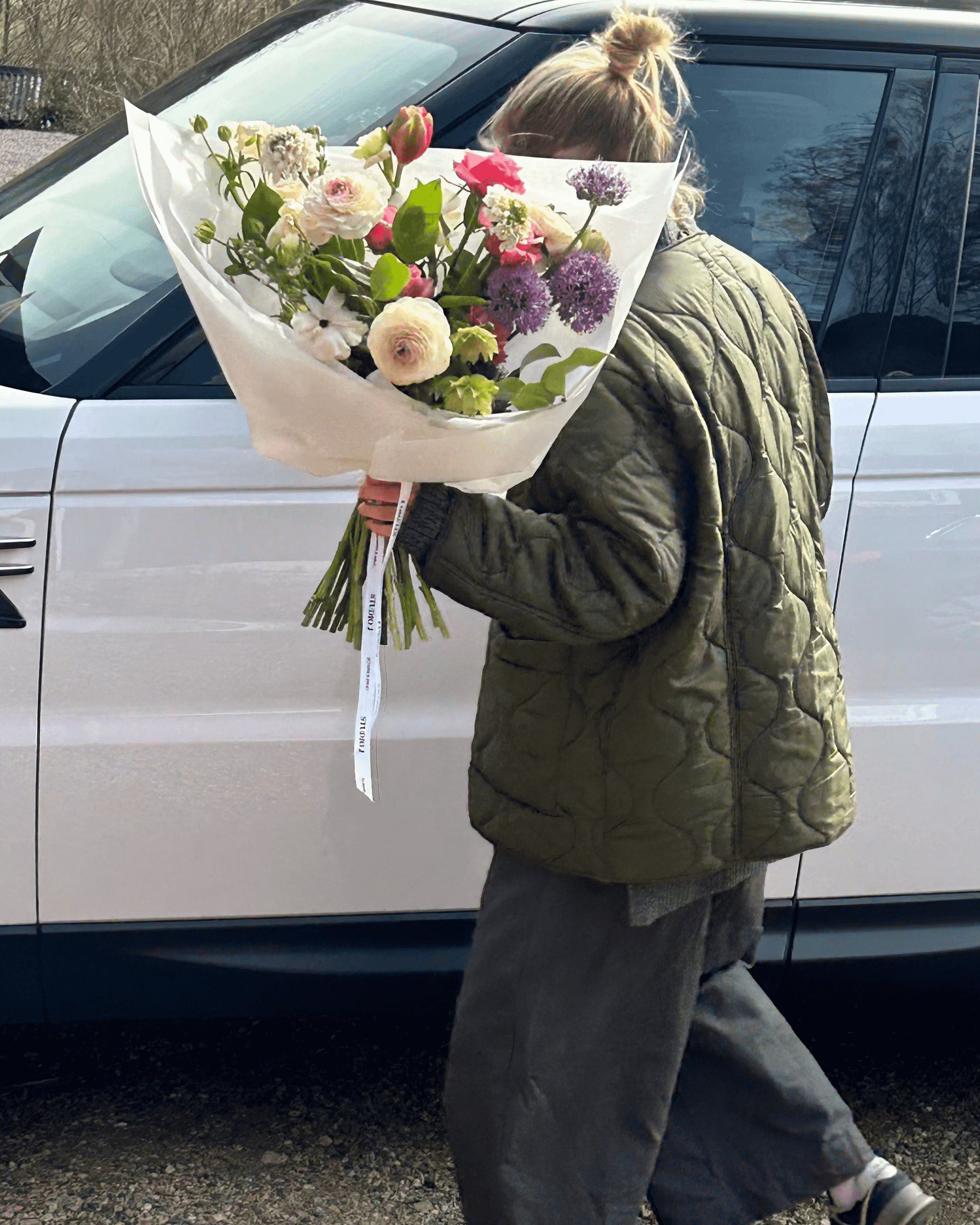 Kvinna i grön jacka som bär en vacker blombukett framför en vit bil